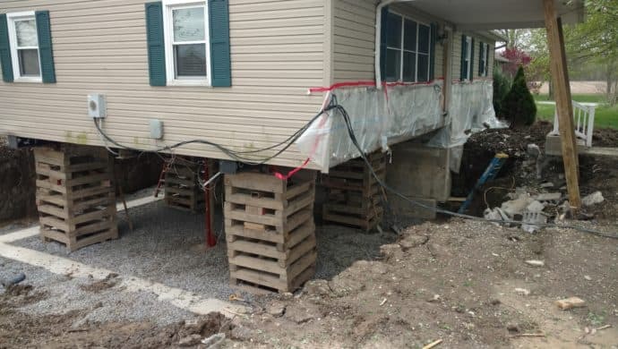 Home elevated on cribbing blocks during foundation wall replacement, showing structural support setup and excavation around the perimeter.