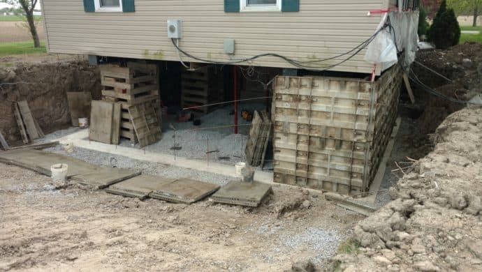 House elevated above ground level while foundation work is underway, with temporary wooden supports and concrete forms in place during excavation.
